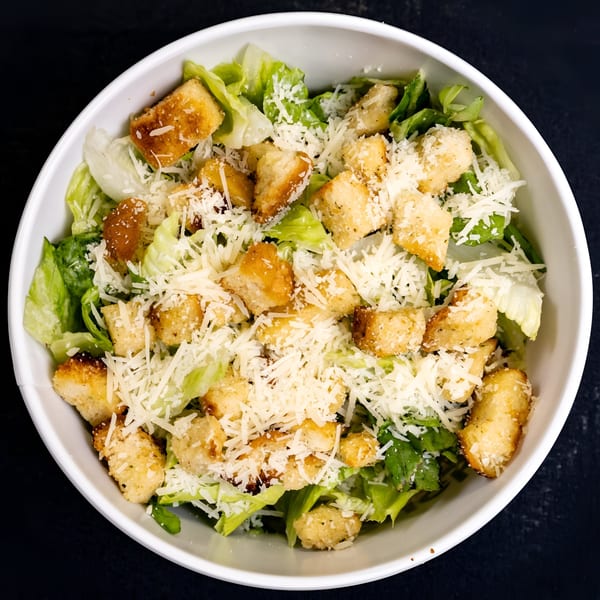 A white bowl filled with fresh green lettuce, golden croutons, and shredded Parmesan cheese.